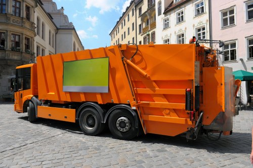 An orange commercial waste collection vehicle parked on a cobblestone street surrounded by historic white and beige buildings with ornate facades and balconies. The truck has a large, rectangular, reflective green panel on its side, a hydraulic arm at the rear for loading or unloading rubbish, and is positioned with its front facing to the left of the image. The environment suggests an urban area, possibly within a town centre, with a clear sky overhead. The scene appears to be part of a rubbish or waste removal operation, characteristic of services provided by companies like Commercial Waste Harpenden, supporting waste disposal or rubbish collection in residential or commercial areas of the region, such as nearby Harpenden.