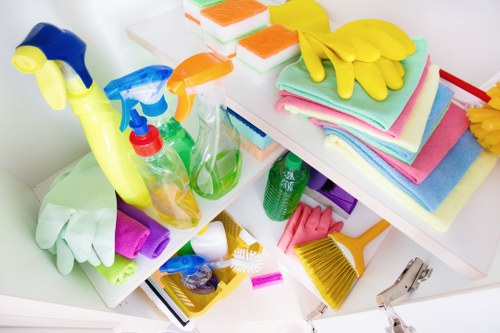 A collection of cleaning supplies and tools arranged on a white surface, including spray bottles filled with cleaning liquids in transparent, green, and blue containers, some with trigger mechanisms, alongside stacked colorful cleaning cloths and towels in shades of pink, blue, and green. Bright yellow rubber gloves are placed atop the towels, with additional gloves in purple and pink beside the bottles. There is a yellow hand brush with stiff bristles and a small purple dustpan with a handle, indicating a cleaning setup typically used for household or commercial cleaning tasks. The background appears to be part of a tidy, indoor storage area or cleaning corner, with a focus on organized hygiene and maintenance supplies, relevant to waste management and rubbish collection services like those offered by Commercial Waste Harpenden in the Harpenden area.