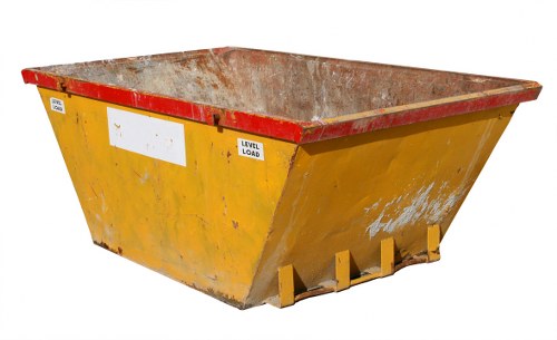 A large, industrial skip positioned on a flat surface, with a rectangular shape and sloped sides. The exterior is painted in a bright yellow colour, but the surface shows visible signs of wear and rust, particularly along the upper edges and corners. The inside of the skip reveals a corroded, reddish-brown metal surface, indicating prior heavy use. At the front bottom edge, there are three yellow metal lifting points or prongs, used for loading and unloading the skip. The background is plain and neutral, with no other objects or environmental context visible, emphasizing the skip's utilitarian design. This image illustrates typical rubbish or waste container equipment used by commercial waste removal services, such as those offered by Commercial Waste Harpenden in Hertfordshire, aligning with local rubbish collection needs.