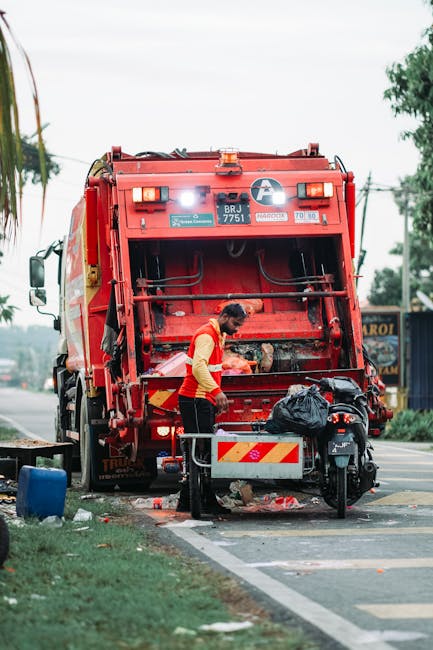 A waste collection worker in a yellow high-visibility vest and red jacket is positioned behind a large red rubbish truck, which is actively engaged in waste pickup on a roadside. The worker is leaning