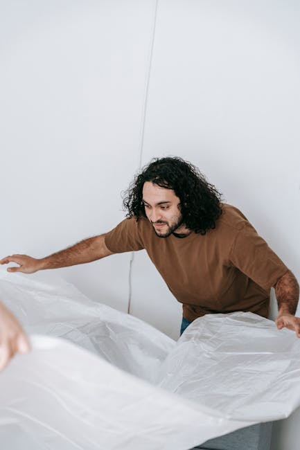 A bedroom interior featuring a large, light-colored upholstered bed with a plain, cream fabric headboard positioned against a beige wall. On either side of the bed, there are matching dark wooden beds